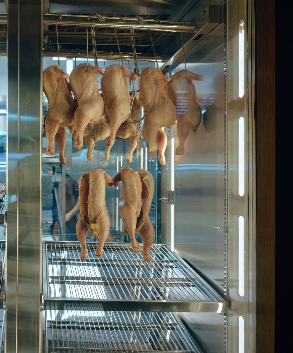 Whole birds and beef cuts hanging in the glass-fronted kitchen chiller at STEKA